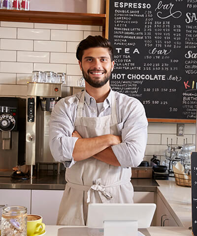 Ventajas para Autónomos - El dueño de una cafetería  con los brazo cruzados sonriendo tras la barra de su negocio Ventajas para Autónomos - El dueño de una cafetería  con los brazo cruzados sonriendo tras la barra de su negocio