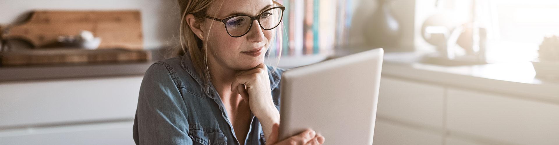 Seguro de Proteccion de Pagos - Mujer con gafas sentada en la cocina de su casa con una tablet en la mano y papeles sobre la mesa, trabajando online