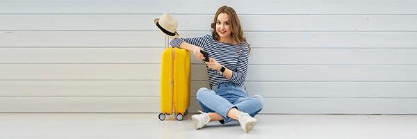Seguro de Asistencia en Viaje - Mujer joven sentada en el suelo con el movil en la mano, junto a su maleta y un sombrero preparando un viaje